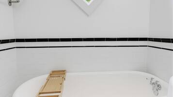 A white freestanding bathtub with a wooden tray, a window with a view of greenery, and black and white patterned floor tiles.