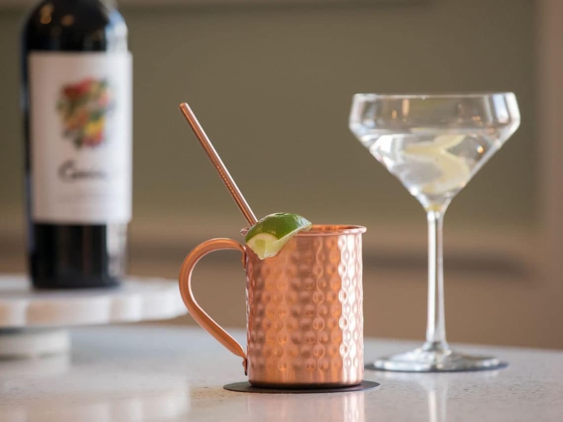A copper mug with a lime wedge and straw, a martini glass, and a bottle of wine on a countertop.