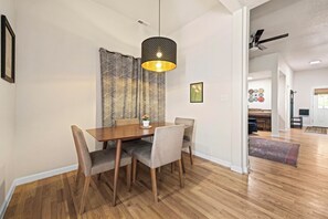 Un coin repas avec une table en bois, des chaises à revêtement gris, un luminaire suspendu et un rideau à motifs.