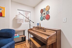 A room with a wooden piano, a blue chair, a window with a view of a brick building, and a wall with decorative plates.