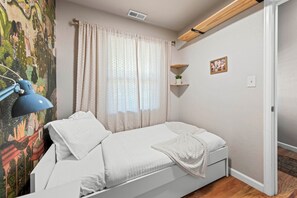 A single bed with a white duvet, a bedside lamp, a window with sheer curtains, a small plant on a shelf, and a framed picture on the wall.