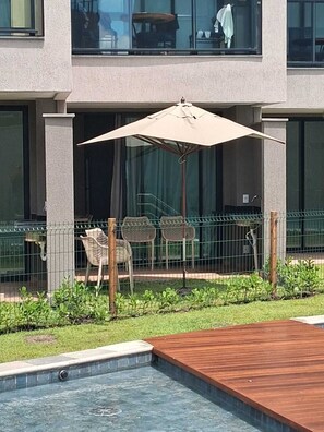A swimming pool with a wooden deck, a metal fence, and a covered seating area with chairs and an umbrella. - F4.009 | Practicality, charm and comfort/Carneiros (Tamandaré)