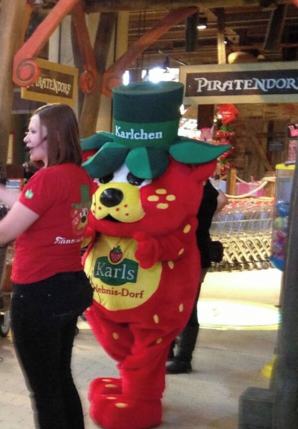 Una persona vestida con un traje rojo y verde de apariencia similar a un personaje publicitario, de pie en una tienda con vigas de madera y un letrero que dice 'Piraten-Dorf'.