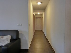 Un pasillo estrecho con una puerta al final, un sofá negro con un cojín y un interruptor de luz en la pared.