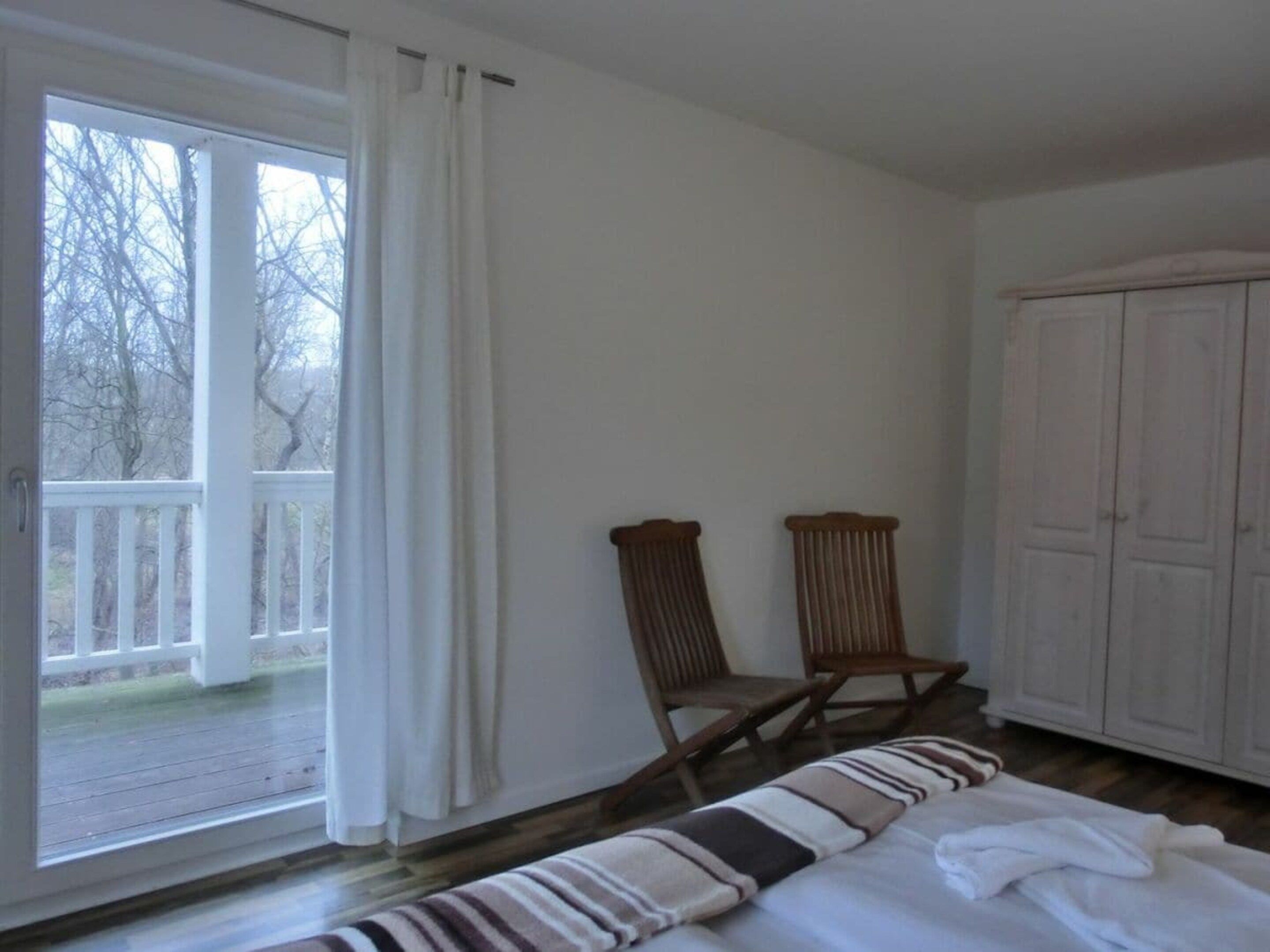 Ein Zimmer mit einem Bett, einem Kleiderschrank, einem Stuhl und Blick auf eine Holzterrasse durch eine Glastür.