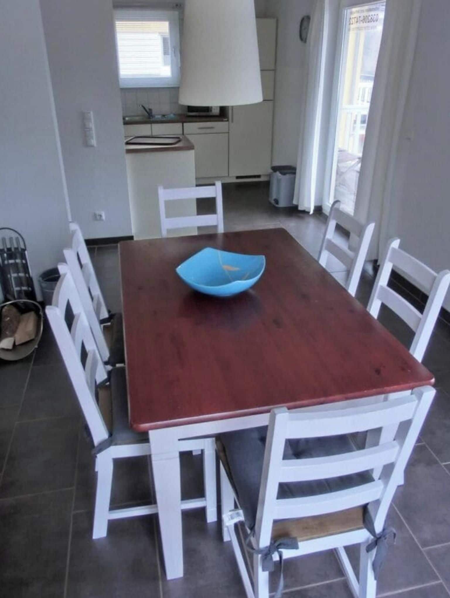 A dining area with a wooden table and white chairs, a blue bowl on the table, and a kitchen in the background.