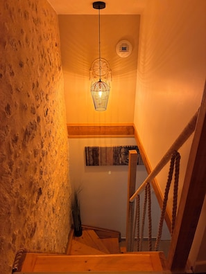 Una escalera con barandilla de madera y cuerda, que lleva a un rellano con un aplique de pared y un cuadro enmarcado.