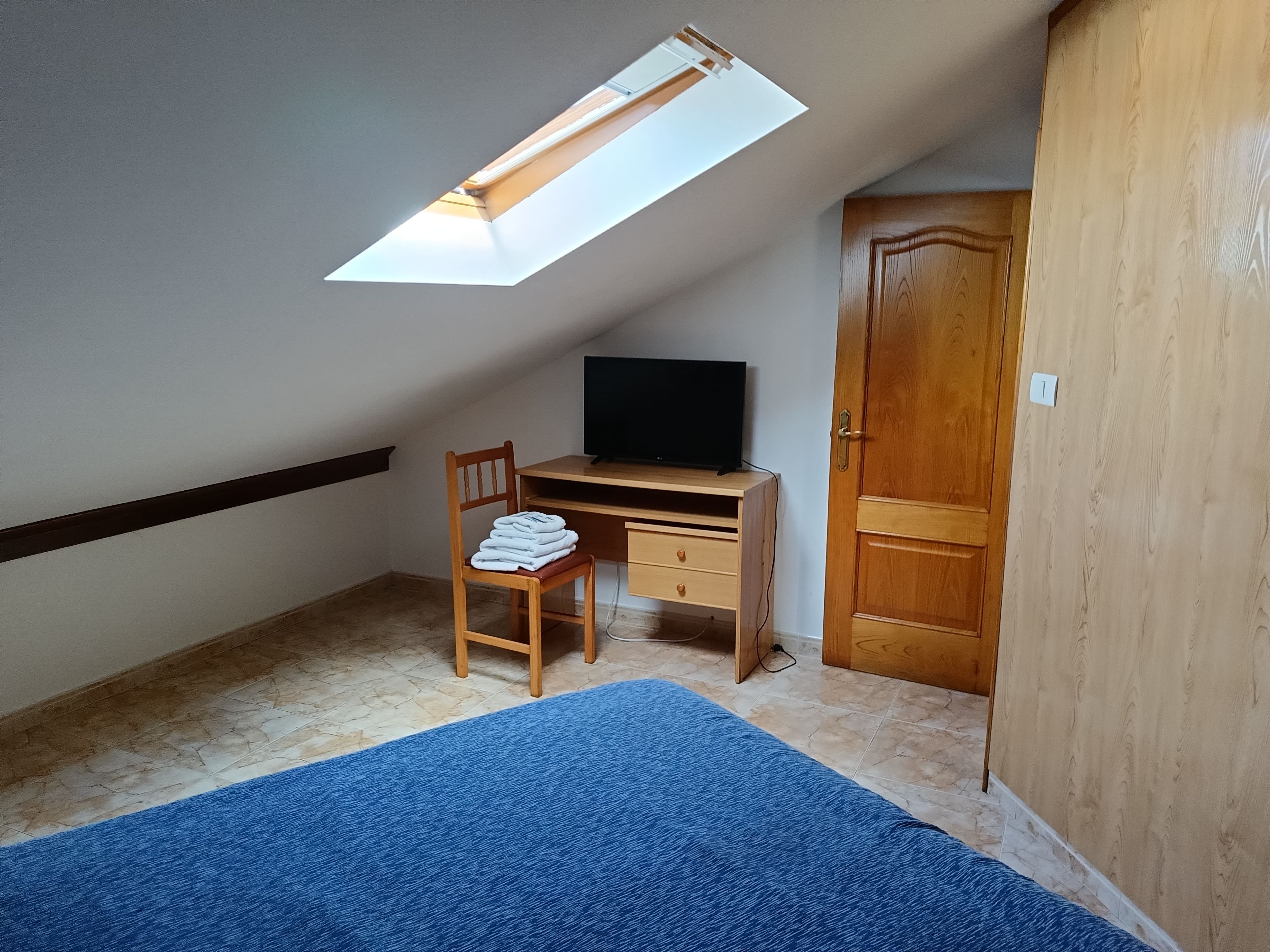 A room with a sloped ceiling, a desk with a computer, a chair, a wardrobe, and a blue carpet.