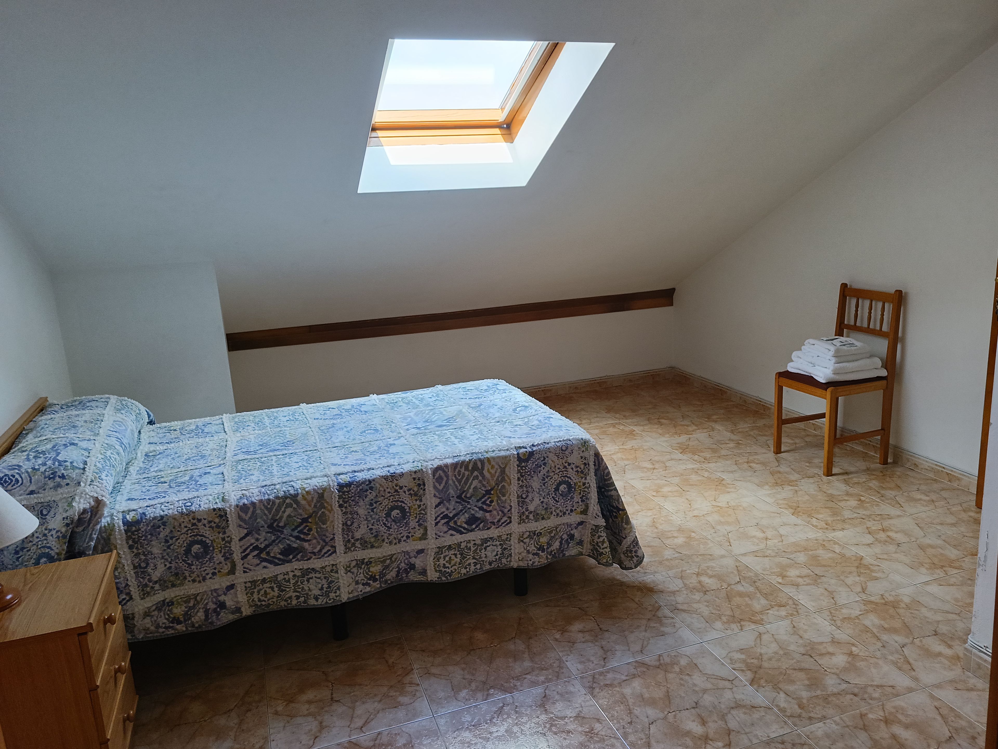 Ein Schlafzimmer mit Dachfenster, einem Bett mit gemusterten Textilien, einem Holzstuhl und einer Kommode.