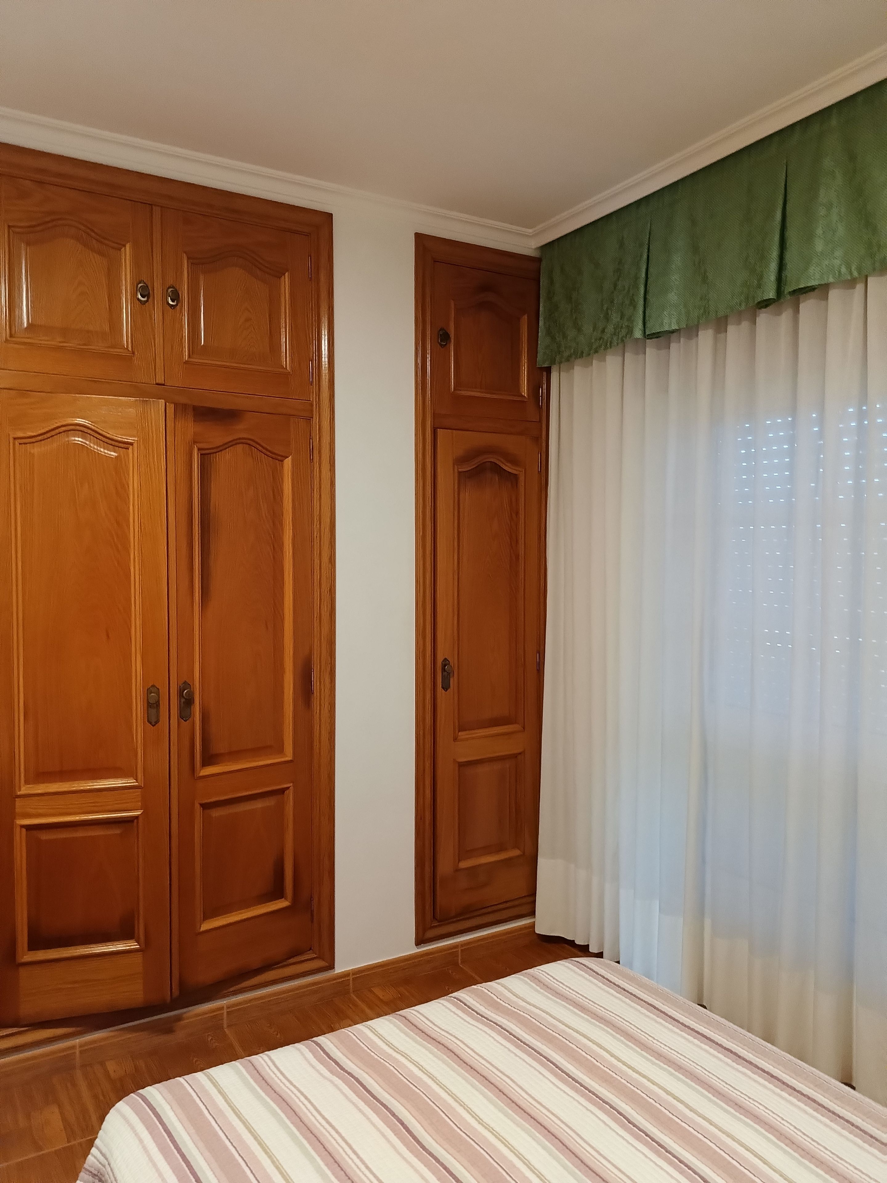 Una habitación con armarios de madera, una cama con cubrecama a rayas y un ventanal con cortinas traslúcidas.