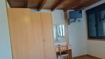 A bedroom with a wooden wardrobe, a desk with a chair, a television mounted on the wall, and a window with blinds.