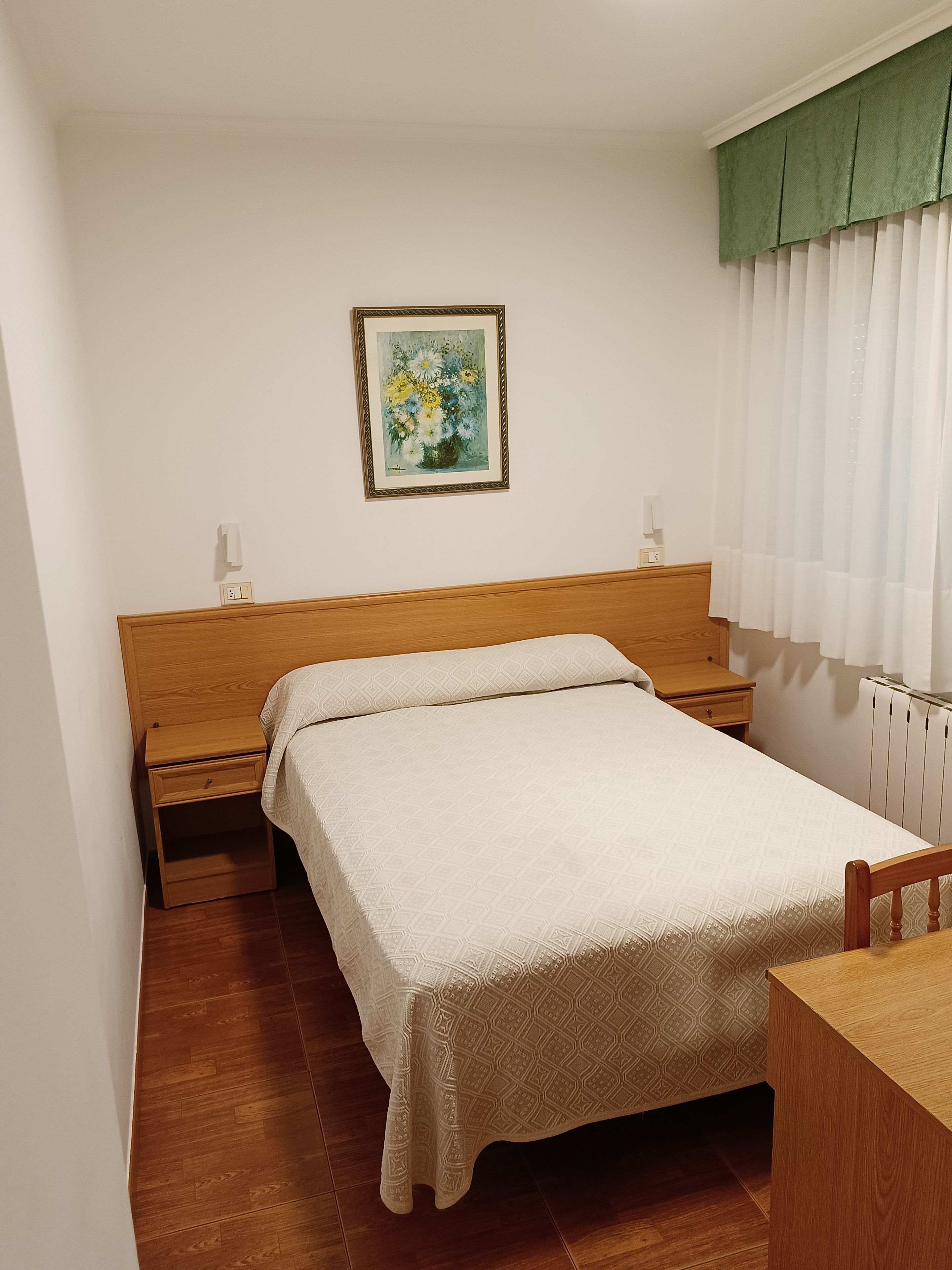 Een enkel bed met houten hoofdbord, nachtkastje en een klein ladeblok. Daarboven hangt een schilderij in een lijst aan de muur.