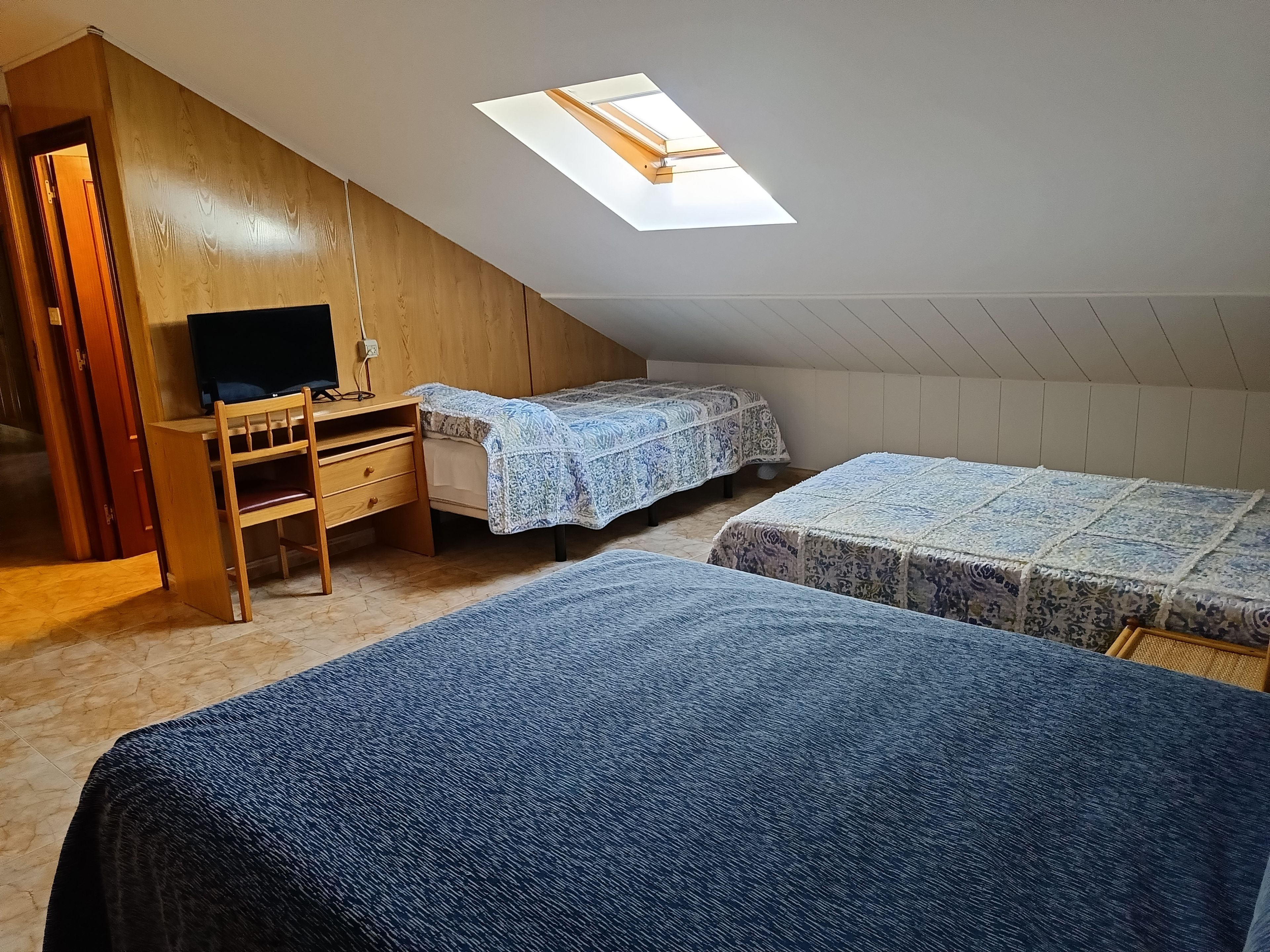 A dormitory room with a sloped ceiling, a skylight, a desk with a computer, two beds with patterned bedding, and a wooden wardrobe.