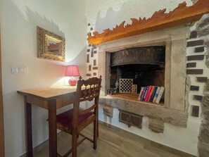 Una habitació amb una llar de foc, una taula de fusta, una cadira, una làmpada i un quadre al mur.