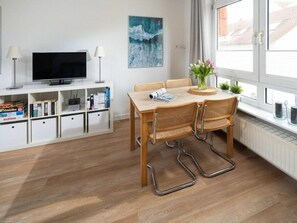A modern living room with a flat-screen TV, white shelving with books, a painting, and a dining area with a table and chairs. - Holiday Apartment in Norderney With Wi-fi (Norderney)