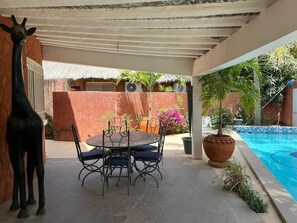 A covered patio with a round table and chairs, a potted plant, and a pool. - Vrbo Property (Saly)