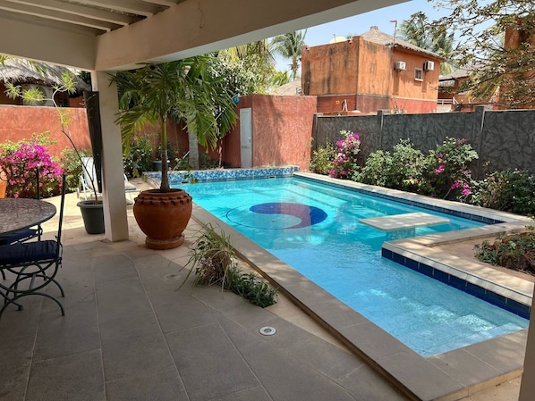 A swimming pool with a tiled deck, a table with chairs, and potted plants. - Vrbo Property (Saly)