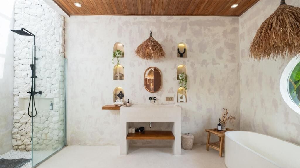 Un baño moderno con pared de piedra, techo de madera, lavabo blanco y un espejo redondo.