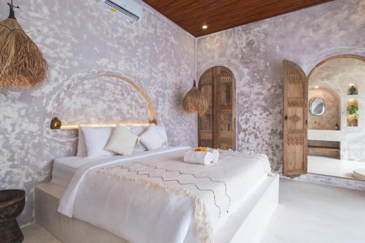 Quarto com cama, luminária pendente, porta de madeira e vista para um banheiro.