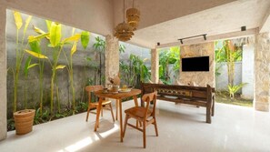 A modern interior with a stone wall, a wooden dining table with chairs, a flat-screen TV mounted on the wall, and a stone fireplace. - Nick Villas (Ubud)