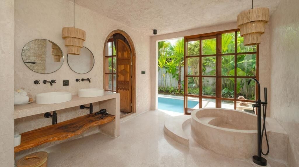 A bathroom with two sinks, a large bathtub, and a window with a view of a pool and greenery.