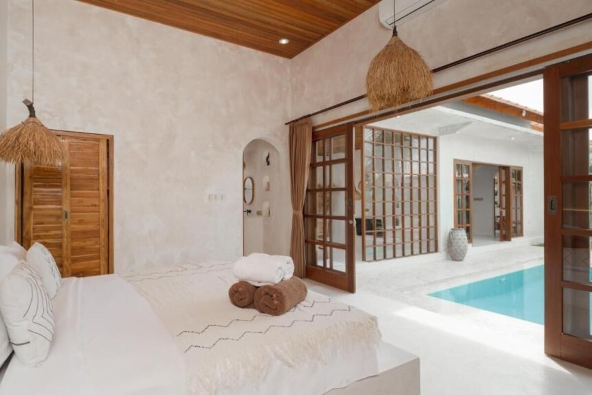 Quarto com cama, porta de madeira, janela e vista para uma piscina ao ar livre.