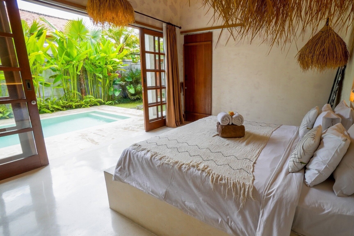 Quarto com cama, almofadas, mesa de cabeceira e vista para uma piscina e vegetação exuberante.