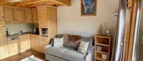 Un salon chaleureux avec un canapé gris, un plancher en bois et une cuisine en arrière-plan.
