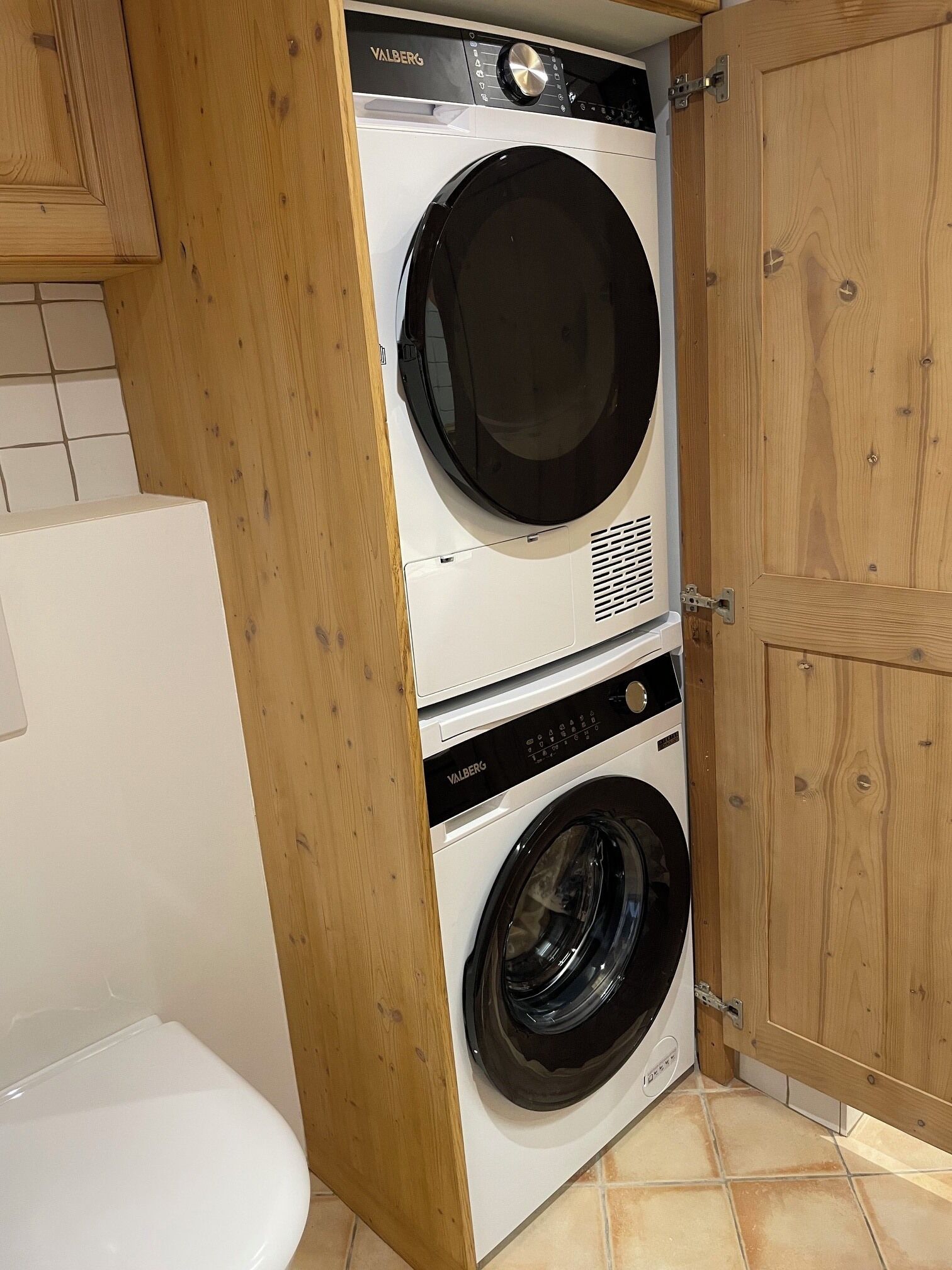 A compact, wall-mounted washer and dryer unit in a bathroom with wooden cabinetry and tiled flooring.