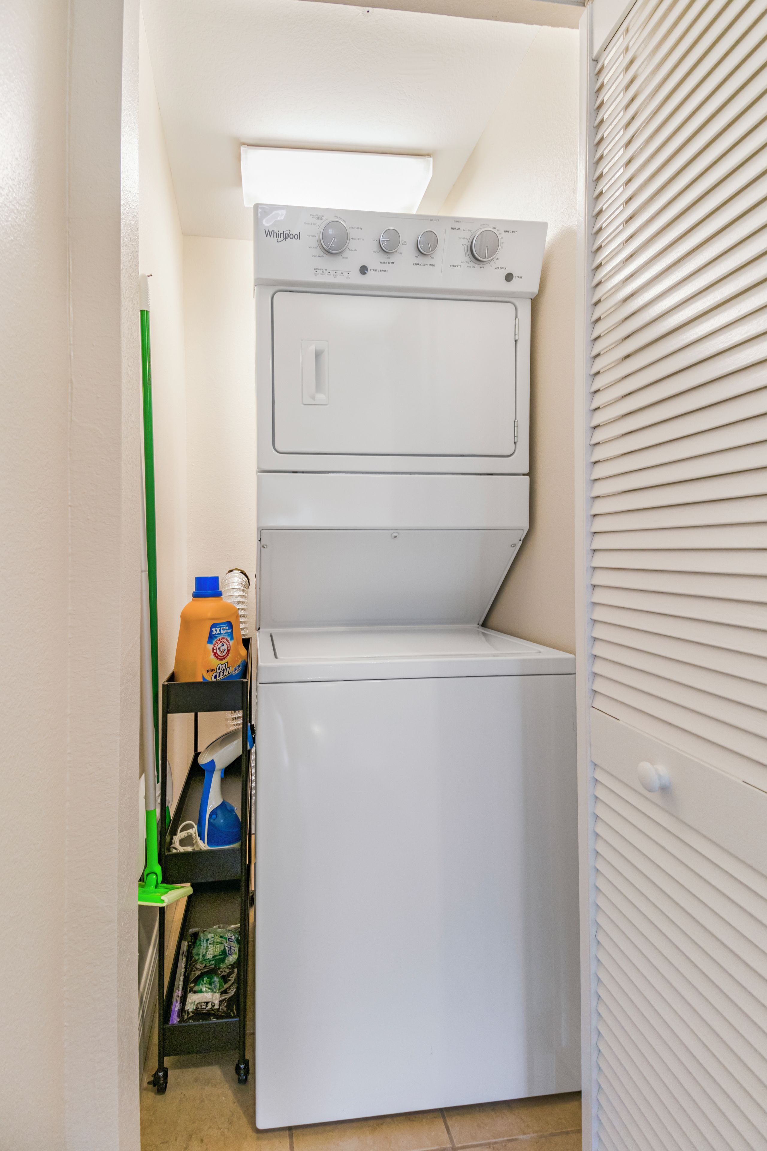 Une petite buanderie équipée d’un lave-linge et d’un sèche-linge à chargement frontal, d’une étagère contenant des produits de nettoyage et d’une serpillière verte.