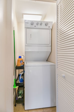 A compact laundry room with a front-loading washer and dryer, a shelf with cleaning supplies, and a green mop.