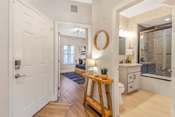 A hallway leading to a bedroom with a bed, a bathroom with a shower, and a small table with a lamp.