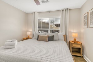 A hotel room with a large bed, two bedside tables with lamps, a window with blinds, and a ceiling fan.