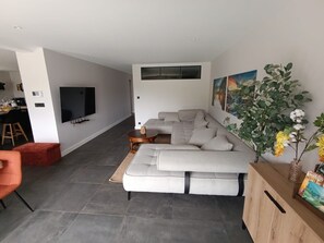 A modern living room with a grey sectional sofa, a flat-screen TV mounted on the wall, and a dining area with a table and chairs.