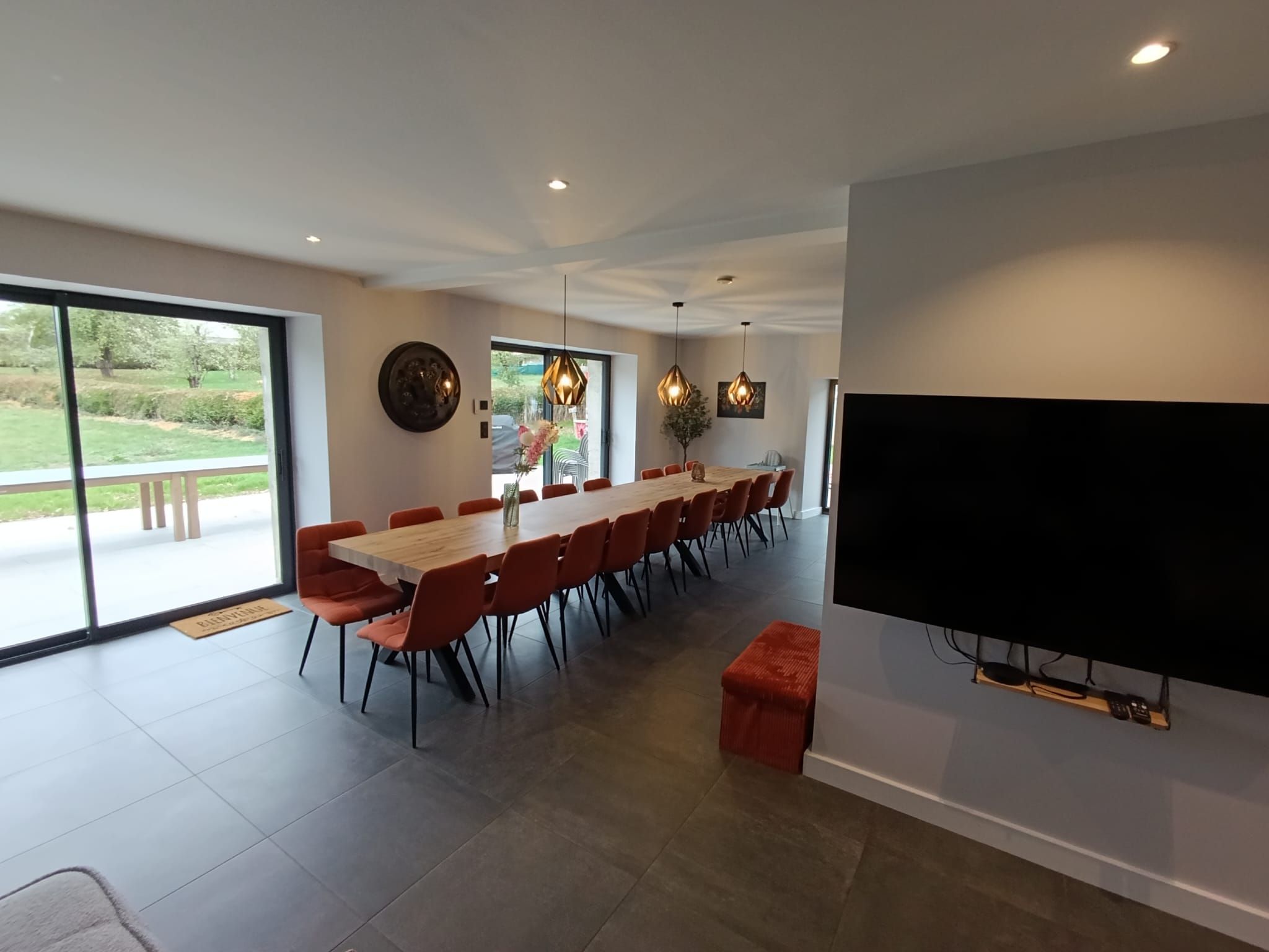 A modern dining area with a long table and orange chairs, a flat-screen TV mounted on the wall, and a large window offering a view of the outdoors.