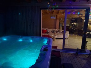 A hot tub with a wooden deck, a glass door leading to a living area with a sofa and a dining table.