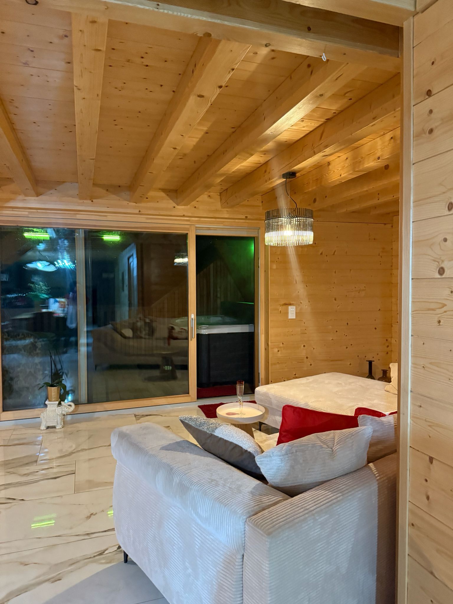 A modern interior with a wooden ceiling, a glass sliding door leading outside, a white sofa with cushions, and a small round table with a glass.