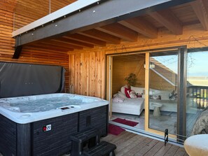 Una terraza de madera con un jacuzzi, un ventanal de vidrio que da a un balcón y un sofá blanco con almohadones.