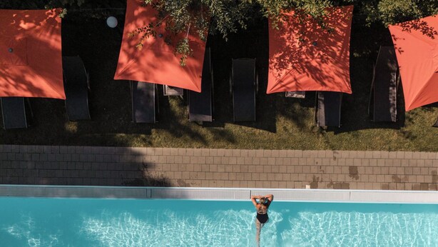 Ein Schwimmbecken mit klarem Wasser, umgeben von einer Ziegelmauer und roten Sonnenschirmen.