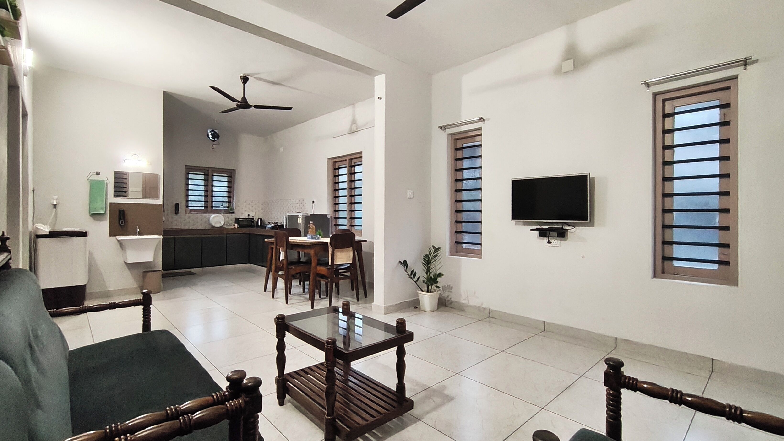 A spacious living area with a dining table, chairs, a sofa, a TV mounted on the wall, and a ceiling fan.