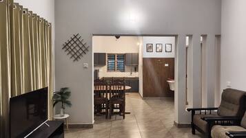 Uma sala de estar moderna com televisão, sofá, área de jantar com mesa e cadeiras, e cozinha visível através de uma porta aberta.