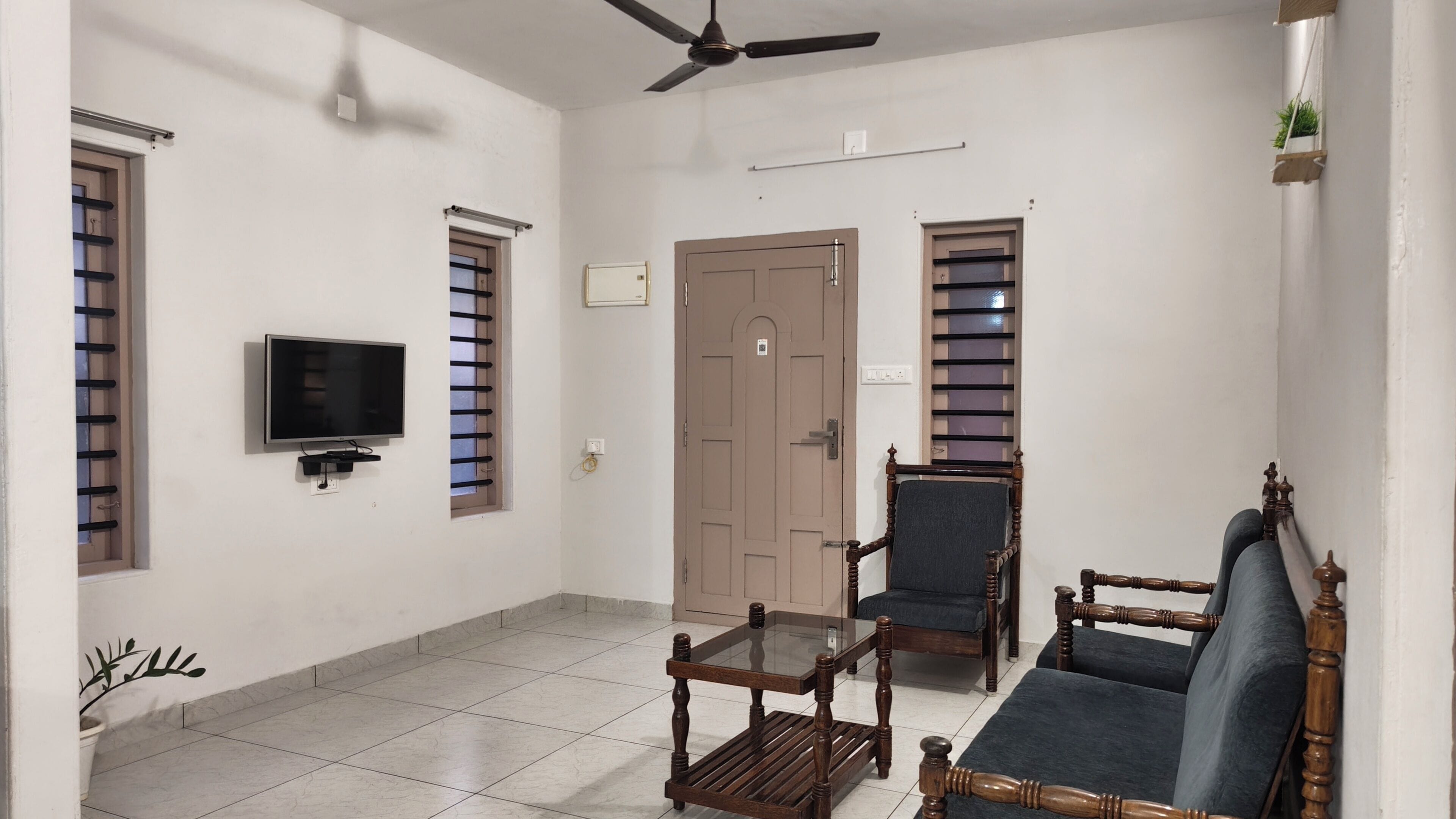 A living room with a television, two wooden chairs, a glass coffee table, a ceiling fan, and a door with a window.