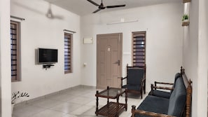 A living room with a television, two wooden chairs, a glass coffee table, a ceiling fan, and a door with a window.