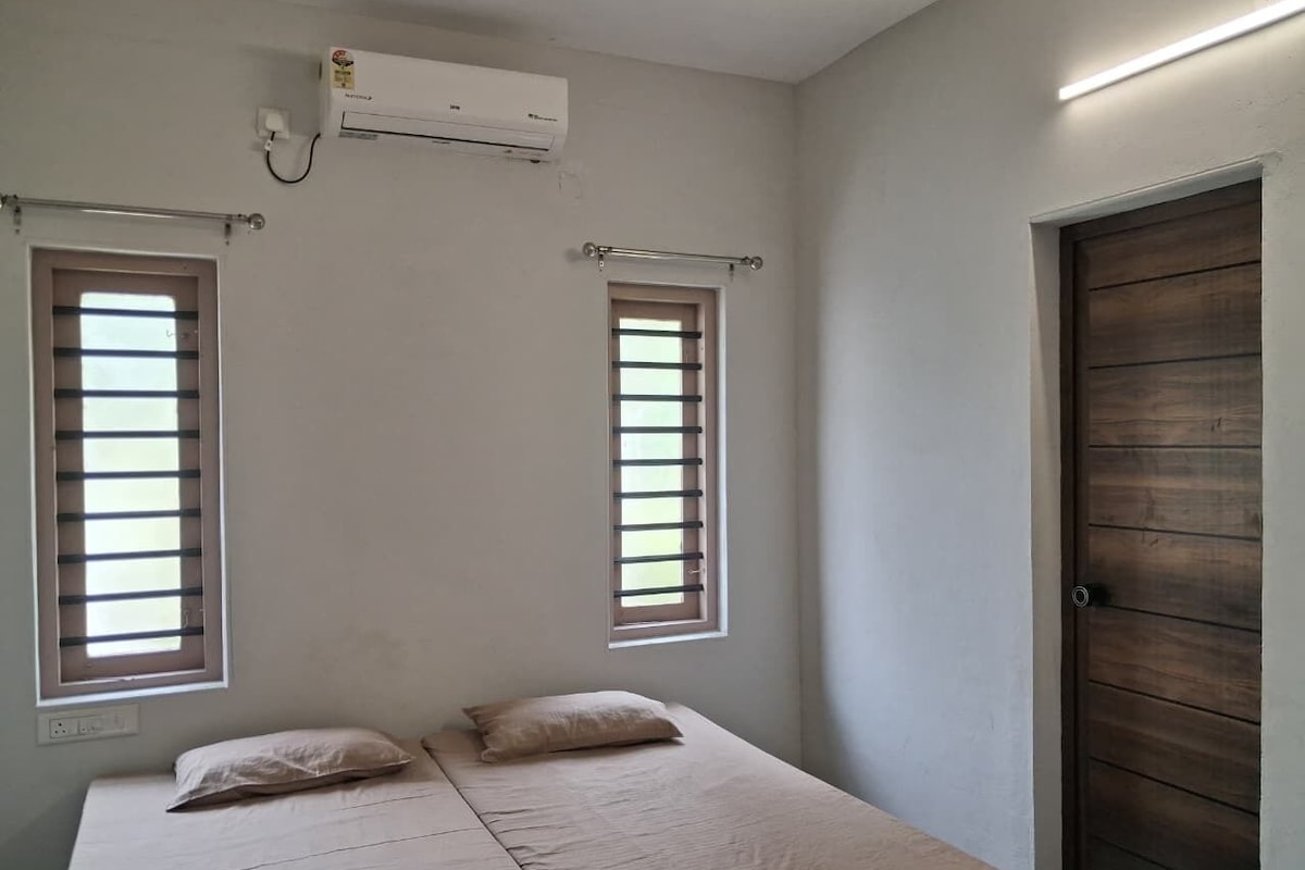 A single bed with two pillows, a window with blinds, a door, and an air conditioning unit.