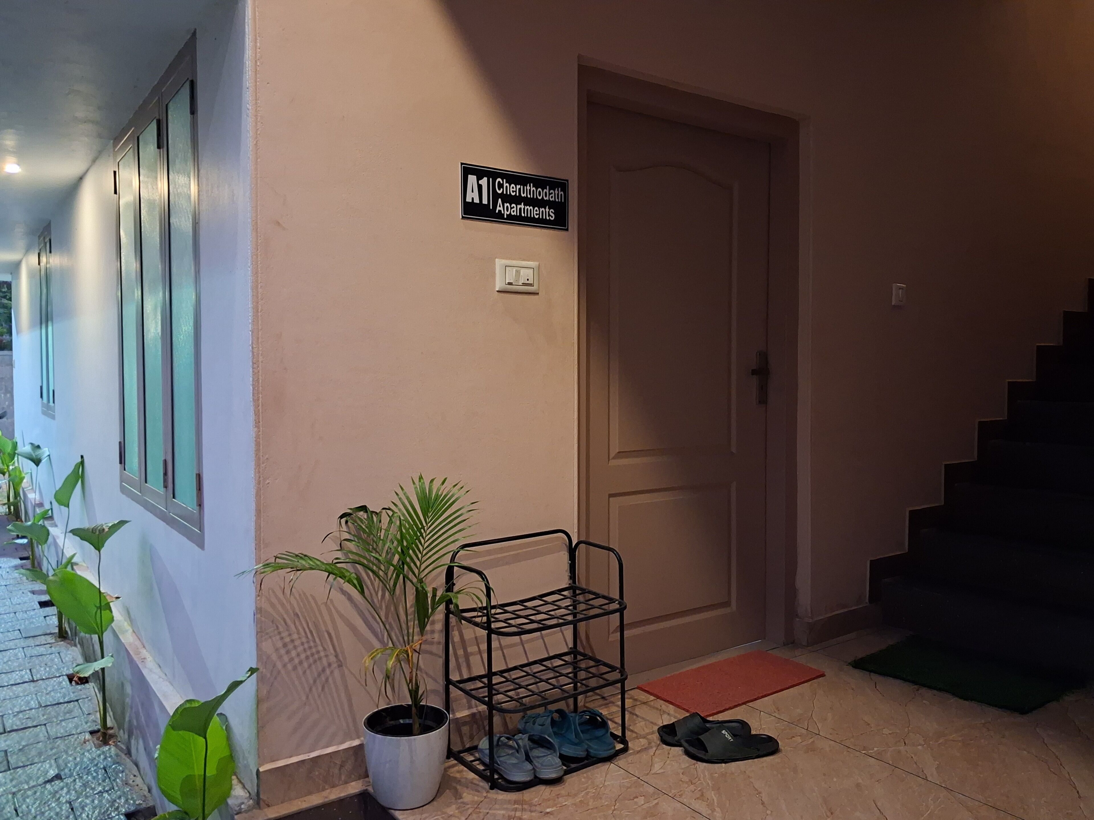 A hallway with a sign for 'A1 Cheruthodath Apartments', a potted plant, a shoe rack, and a door.