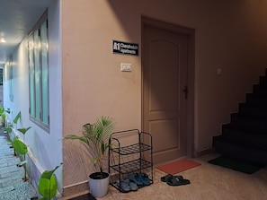 A hallway with a sign for 'A1 Cheruthodath Apartments', a potted plant, a shoe rack, and a door.