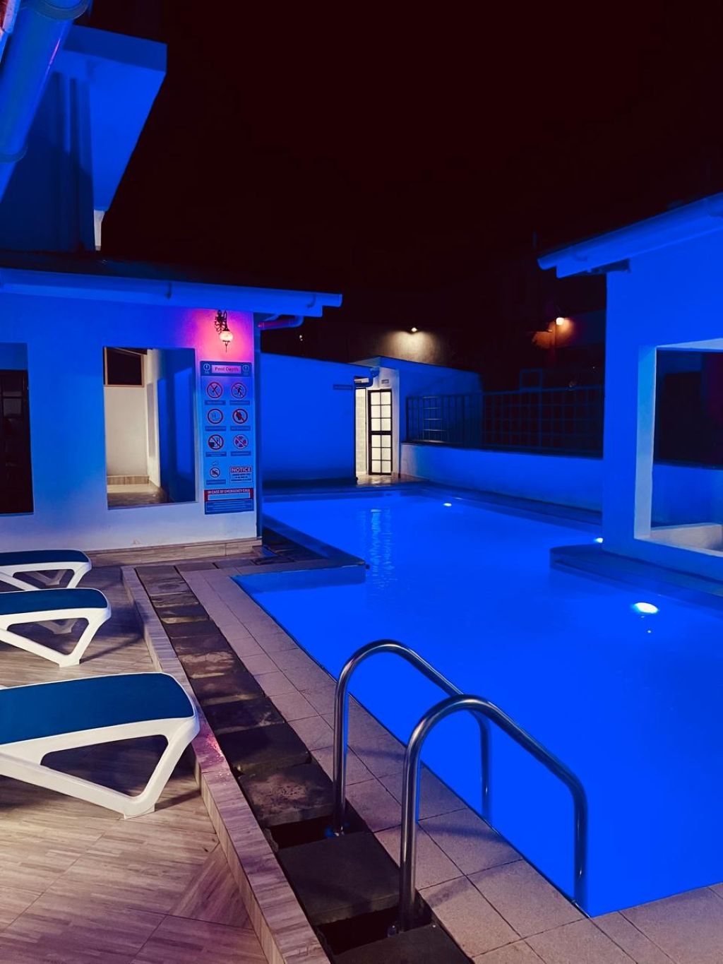 A swimming pool area with a blue-lit exterior, a poolside seating area, and a pool ladder.