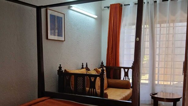 Een hemelbed met een wit-oranje beddeken, een tafeltje met een stoel en een raam met gordijnen.
