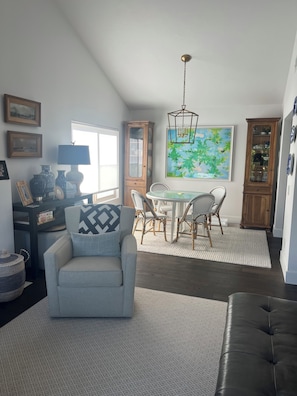 A living room with a grey armchair, a dining table with chairs, a chandelier, and a large painting on the wall. - Beautifully remodeled and furnished Spacious 3 condo in Oaks North 55+ Community (San Diego)
