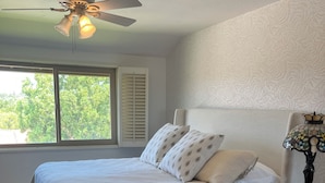 Ein Schlafzimmer mit einem Bett, einem Nachttisch, einer Lampe, einem Deckenventilator und einem Fenster mit Jalousien.